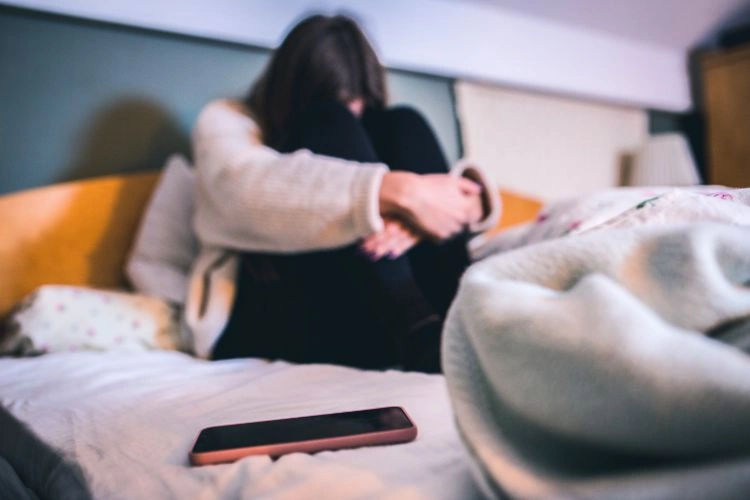 Femme déprimée sur son lit avec son téléphone posé à côté représentant les formes de cyberharcèlement